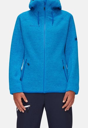 Veste à capuche bleue avec fermeture éclair, avec poches latérales zippées, portée par une personne en pantalon foncé avec une poche zippée sur la cuisse.