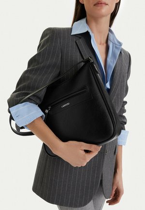 Mujer con blazer gris de rayas y camisa azul sosteniendo un bolso de hombro negro de cuero LASOCKI sobre fondo blanco.