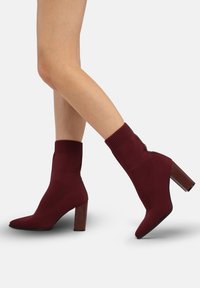Botines color burdeos hechos de material de punto, con punta puntiaguda y tacón grueso rectangular, con un puño acanalado en la parte superior.