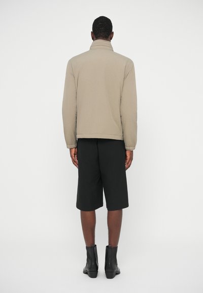 Jachetă bej căptușită cu guler înalt, purtată cu pantaloni scurți negri. Jacheta are o textură netedă și manșete elastice, creând o croială lejeră.