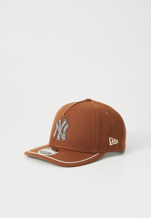 Cappellino da baseball marrone in tessuto, con un logo ricamato in evidenza sulla parte frontale, visiera curva e rifiniture decorative.