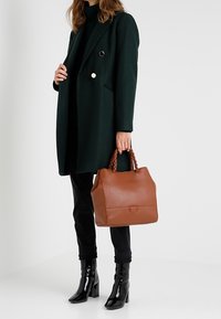 Bruine leren handtas met een gevlochten handvat, gecombineerd met een donkergroene jas, zwarte skinny jeans en glanzende zwarte enkellaarzen.