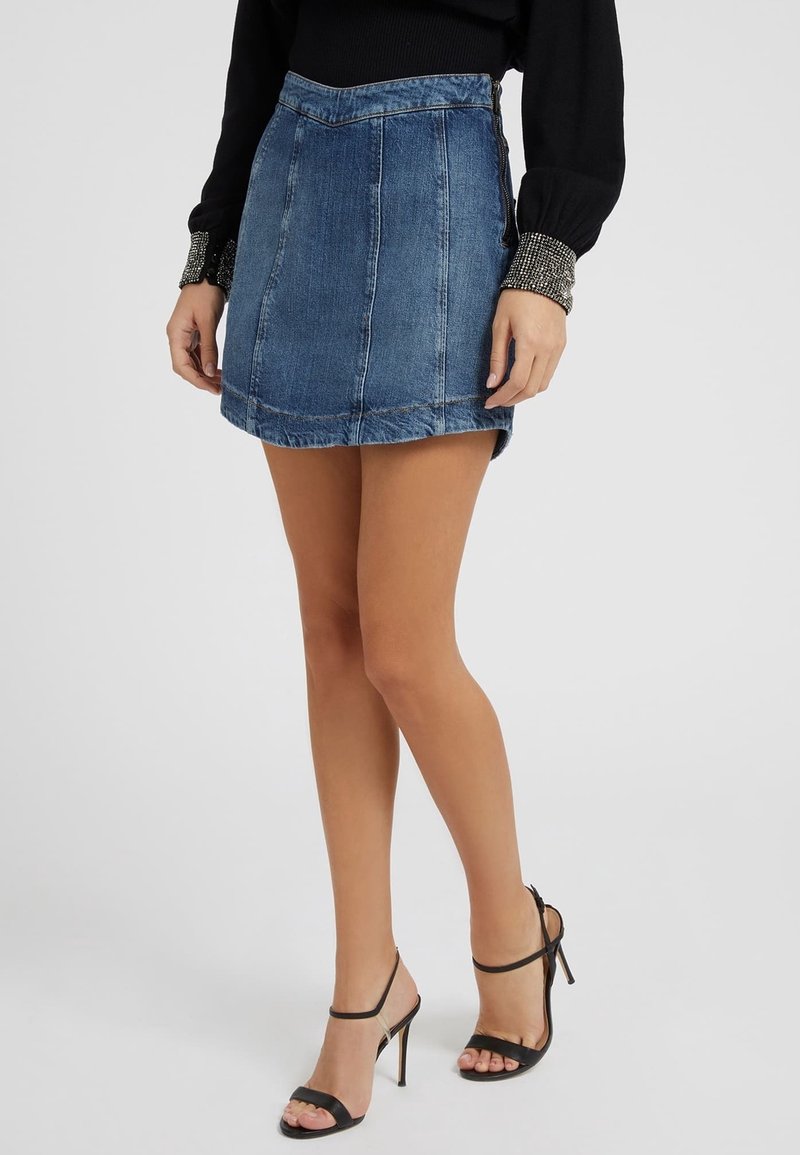 Mini-jupe en denim taille haute, de couleur bleu délavé, avec des détails de couture visibles. Associée à des sandales à talons hauts noires à lanières.