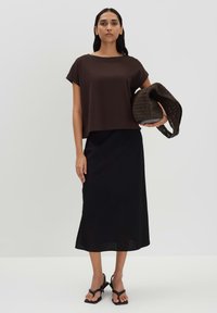 Braunes T-Shirt mit kurzen Ärmeln, kombiniert mit einem schwarzen Rock. Das Outfit zeichnet sich durch eine lockere Passform, glatte Textur und schwarze Riemchensandalen aus.