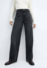 Mustat leveälahkeiset farkut, joissa korkea vyötärö, etutaskut ja napillinen kiinnitys, yhdistettynä kermaan vivahtavaan ribbineulepaitaan ja tummiin kenkiin.