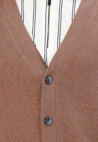 Hnědý pletený cardigan se dvěma černými knoflíky přes bílou košili s vertikálními černými pruhy a předním zipem.