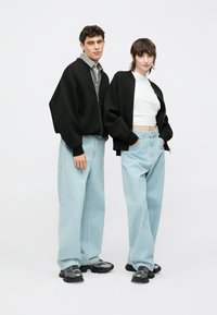 Twee personen staan naast elkaar. De een draagt een zwarte bomberjack, een gestreept shirt en lichtblauwe wijde spijkerbroek. De ander draagt een kort wit topje en een zwarte jas. Beiden dragen stevige zwarte sneakers.