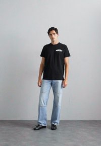 Schwarzes Baumwoll-T-Shirt mit weißem "Ducati"-Logo, kombiniert mit hellblauen Jeans mit gesteppten Nähten und schwarzen Schuhen.