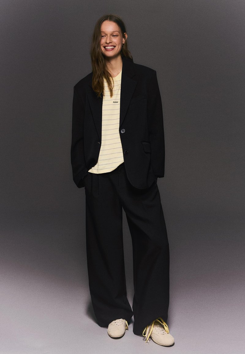 Femme souriante portant un blazer noir surdimensionné et un pantalon large noir par-dessus un t-shirt beige rayé avec des baskets beiges, debout contre un fond gris.