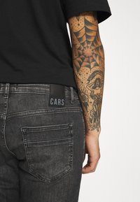 Zwarte T-shirt gecombineerd met donkergrijze spijkerbroek, met een zwart label met "CARS". Zichtbare armtatoeage met ingewikkelde ontwerpen en schaduwwerking.