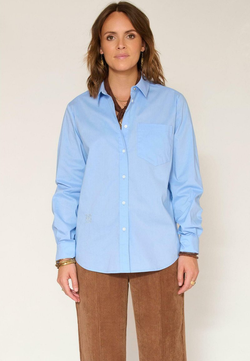 Chemise bleu clair à manches longues et poche poitrine, avec un détail brodé discret. Associée à un pantalon en velours côtelé marron.