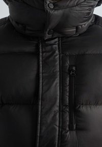 Veste matelassée noire avec un col montant, dotée d'une poche zippée à l'avant, d'une texture matelassée et d'une fermeture à boutons. Tissu lisse et résistant à l'eau.