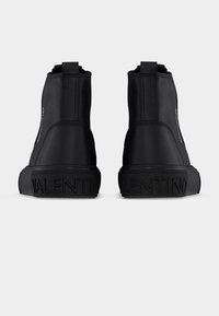 Μαύρα μποτάκια αστραγάλου από λείο ύφασμα, με χοντρή λαστιχένια σόλα και ανάγλυφο όνομα του бренδου στη φτέρνα. Απλός, μινιμαλιστικός σχεδιασμός.