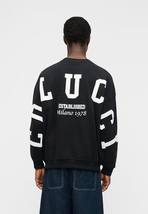 Homme aux cheveux courts et bouclés portant un sweat-shirt noir surdimensionné avec de grandes lettres blanches L U C C I dans le dos et sur les manches, ainsi que le texte "Established Milano 1978".