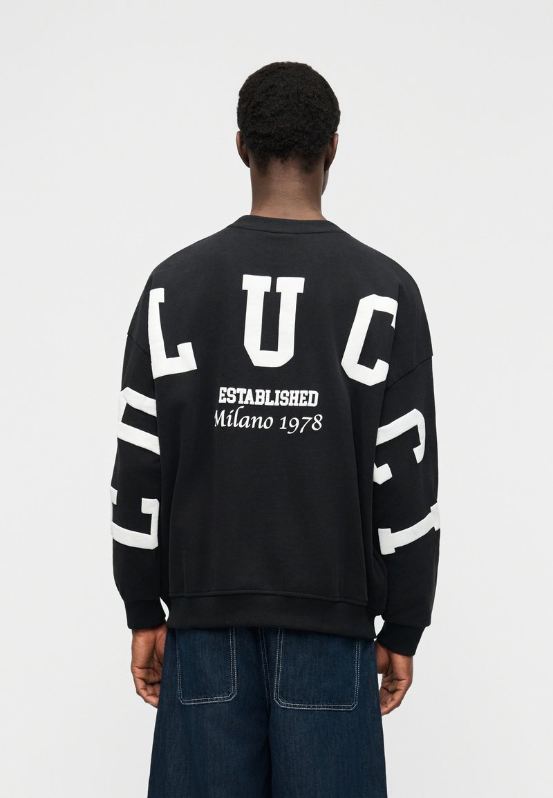 Homme aux cheveux courts et bouclés portant un sweat-shirt noir surdimensionné avec de grandes lettres blanches L U C C I dans le dos et sur les manches, ainsi que le texte "Established Milano 1978".