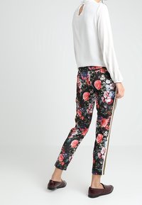 Vit blus med långa ärmar och nyckelhål baktill, parat med svarta blommiga byxor med röda och rosa rosor samt gyllene sidostråkar. Bruna skor.