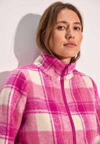 Veste en polaire à motif à carreaux rose et blanc, avec un col haut et une fermeture éclair à l'avant. Texture douce avec un contraste de couleurs vif et un design carré.