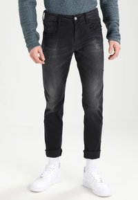 Jeans de denim negro con un corte ajustado, textura sutilmente desgastada, diseño de cinco bolsillos y dobladillos enrollados, combinados con zapatillas blancas y calcetines de caña corta.