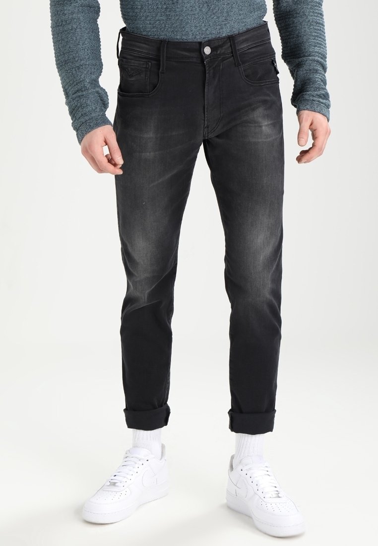 Jeans de denim negro con un corte ajustado, textura sutilmente desgastada, diseño de cinco bolsillos y dobladillos enrollados, combinados con zapatillas blancas y calcetines de caña corta.