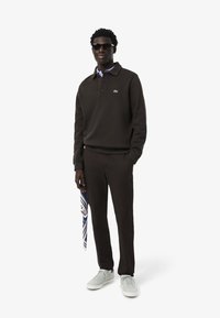 Donkerbruine polo sweatshirt, gedragen over een lichtblauw overhemd met kraag, gecombineerd met bijpassende broek en lichtgrijze sneakers. Houdt een geprijsde sjaal vast.