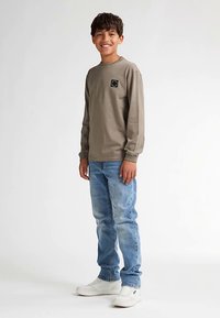 Pitkähihainen oliivinvihreä paita, jossa on pieni musta logo rinnassa, yhdistetty vaalean sinisiin farkkuihin ja valkoisiin tennareihin.