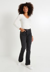 Mujer con cabello rizado, vistiendo una blusa blanca de manga larga, jeans negros de cintura alta y zapatillas blancas, de pie con una mano en el bolsillo.