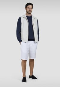 Giacca senza maniche grigio chiaro sopra un maglione blu navy, pantaloni corti bianchi e scarpe slip-on nere. Design semplice con zip frontale e tasche.