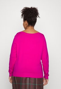 Suéter rosa brillante con mangas largas, un escote amplio y un dobladillo acanalado. Llevado sobre una falda marrón a cuadros con líneas moradas y verdes.