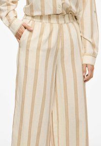 Gaišbēša krāsas platajās biksēs ar oranžām svītrām. Tām ir elastīga josta un sānu kabatas. Audums šķiet viegls un mīksts.