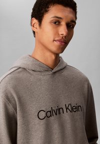 Ung mann med kort krøllete hår som har på seg en grå hettegenser med "Calvin Klein"-logo på brystet mot en ensfarget bakgrunn.