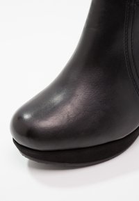 Gros plan sur l'avant d'une chaussure en cuir noir avec une texture lisse et des détails de couture sur le côté, posée sur une surface blanche.