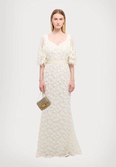 Witte bloemenkant jurk met doorzichtige pofmouwen en een satijnen riem. Model houdt een kleine metalen tas vast. Glad, aansluitend silhouet tot op de vloer.