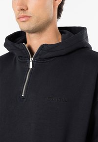 Svart hettegenser med glidelås laget av mykt stoff, med høy krage, sølvfarget glidelås og preget logo på fremsiden. Enkel og minimalistisk design.