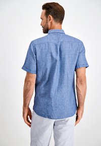 Camicia di lino blu a maniche corte con colletto, cuciture visibili e una trama sottile; abbinata a pantaloni corti leggeri a righe.