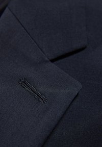 Navy blauwe stof close-up die een gestructureerde weving toont, met een zichtbare op maat gemaakte revers en gestikte accentdetails op het zakgebied.