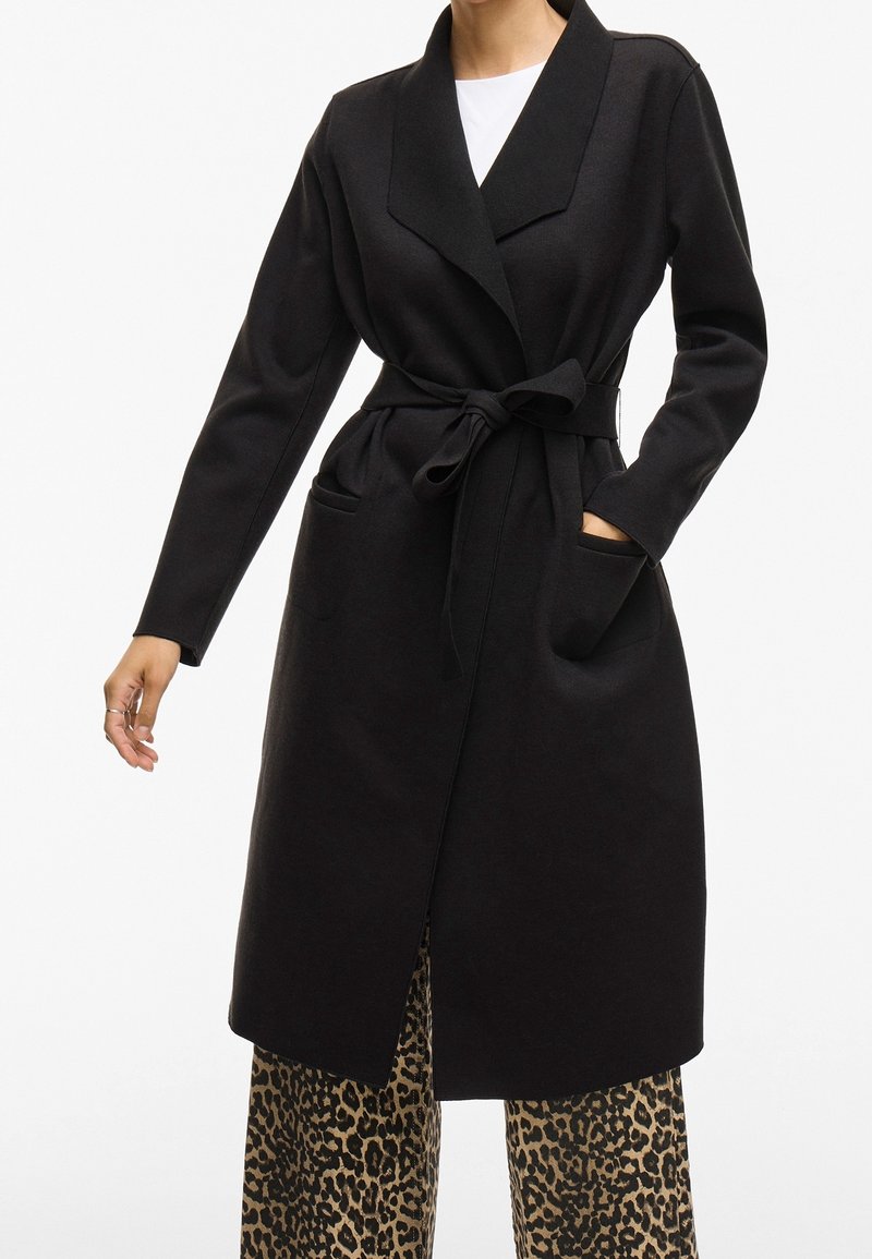Personne portant un long manteau noir ceinturé par-dessus un haut blanc et un pantalon large à imprimé léopard, avec une main dans la poche du manteau.