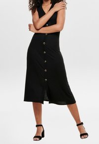 Femme portant une robe midi noire sans manches avec des boutons à l'avant et des talons noirs ouverts à la cheville, debout les bras croisés.