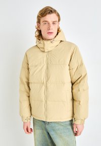 Beige puffer jacket with a zippered front, drawstring hood, and quilted texture. Features two side pockets and elastic cuffs.