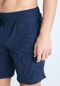 Marine shorts med elastisk midjebånd, snor, og fremtredende brodert "BOSS"-logo i kontrastfarge langs siden.