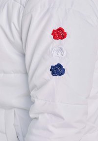 Manica di giacca imbottita bianca con tre rose ricamate: rossa, bianca e blu, disposte verticalmente, con una texture liscia e polsino elastico.