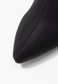 Chaussure en tissu noir avec un design à bout pointu, texture lisse et couture visible sur le côté.