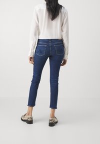 Jeans de mezclilla azul oscuro con un corte ajustado, que cuentan con bolsillos traseros y un diseño bordado sutil. Combinados con una blusa ligera de manga larga y zapatos con estampado de serpiente.