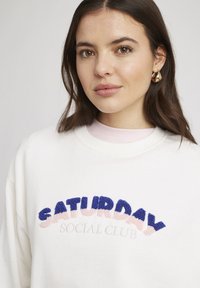 Sweatshirt blanc avec "SATURDAY SOCIAL CLUB" en broderie texturée bleue et rose. Encolure ras du cou et superposé avec une chemise rose clair en dessous.