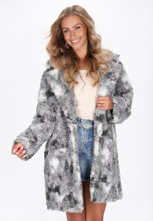 Femme souriante avec de longs cheveux ondulés portant un manteau texturé gris et blanc sur un haut beige et une jupe en denim avec une ceinture décorative.