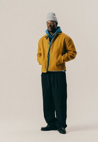 Άνδρας που στέκεται με τα χέρια στις τσέπες, φοράει γκρι πλεκτό σκουφί, γυαλιά, μπλε φούτερ, μουσταρδί μπουφάν, μαύρο φαρδύ παντελόνι και μαύρα παπούτσια σε ανοιχτό φόντο.