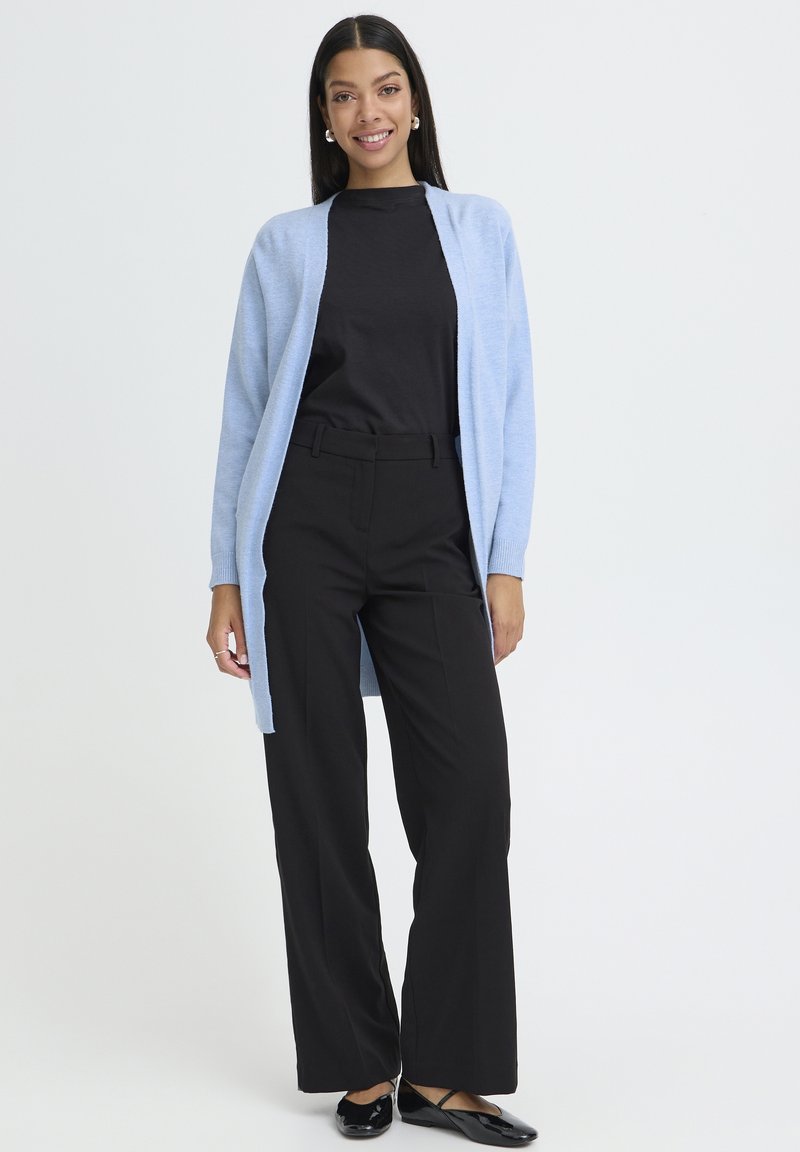 Lyseblå lang cardigan over en sort top, sat sammen med sorte vidde bukser og sorte flade sko. Glat tekstur og afslappet pasform.