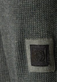 Grüner Strickstoff mit einem strukturierten Muster; enthält ein dunkles quadratisches Patch mit geprägtem rundem Logo und hellgrauem Rand.
