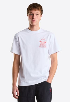 Młody mężczyzna w białej koszulce z krótkim rękawem z czerwonym logo góry i napisem „The North Face”, w czarnych spodenkach, stojący z jedną ręką w kieszeni.