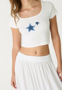 Hvid cropped t-shirt lavet af blødt stof, med to broderede blå stjerner. Sammen med en svajende hvid nederdel.