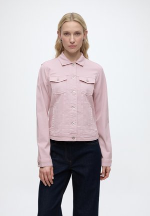 Frau trägt eine hellrosa Jacke mit Knöpfen und Klappentaschen über dunkelblauen Jeans, steht vor einem schlichten weißen Hintergrund.